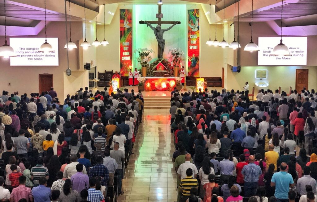 A full Catholic church during Mass, the congregation seated under pendant lights, a large crucifix and red banners behind the altar