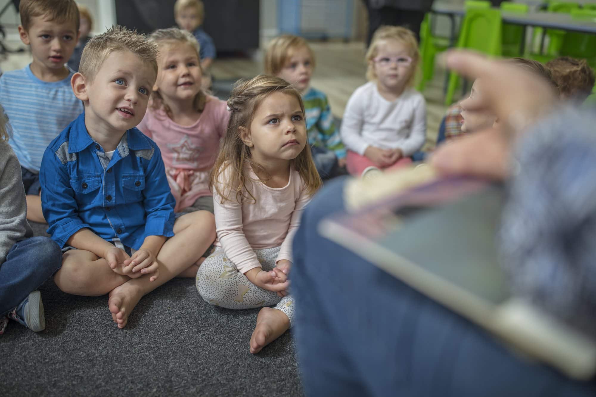 Six small children sit cross-legged on a grey carpet, looking up at an adult reading a picture book that is blurred in the foreground