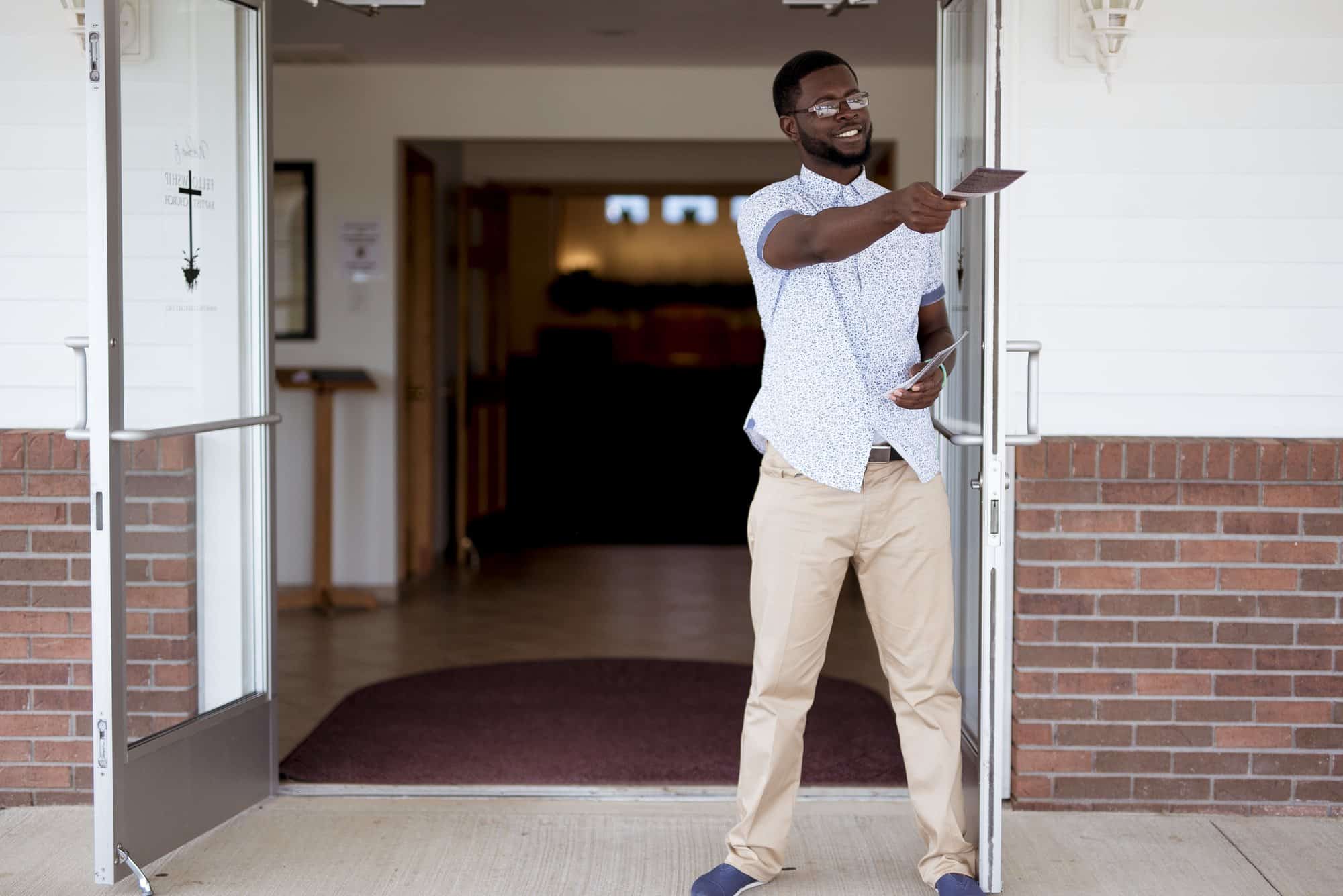 A smiling young man in a patterned short-sleeved shirt and khaki trousers stands in the open glass doors of a brick church, holding out a leaflet to someone arriving