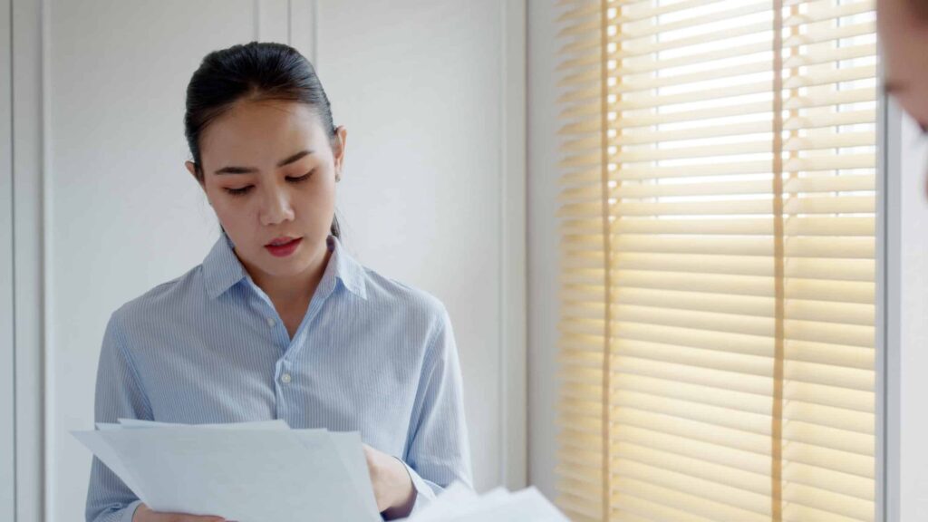 A woman in a striped shirt reading a sheaf of papers beside a window with wooden blinds