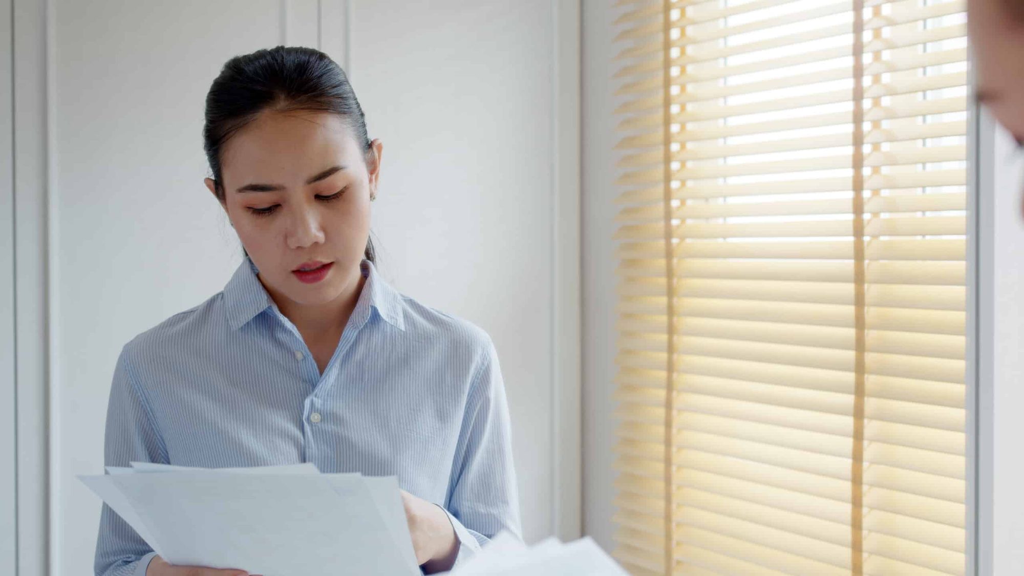 A woman in a striped shirt reading a sheaf of papers beside a window with wooden blinds