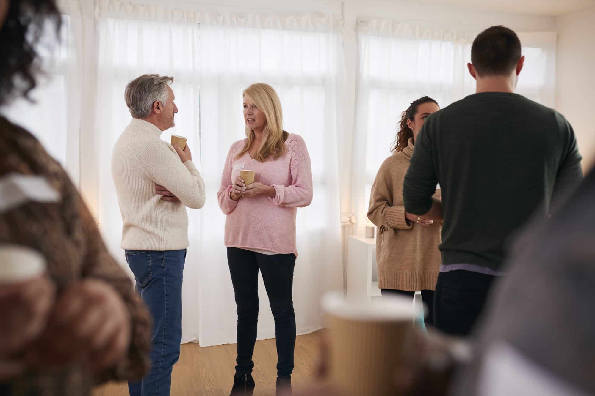 A man in a cream jumper and a woman in a pink jumper talk over paper cups of coffee in a bright room with white curtains, with other people chatting around them