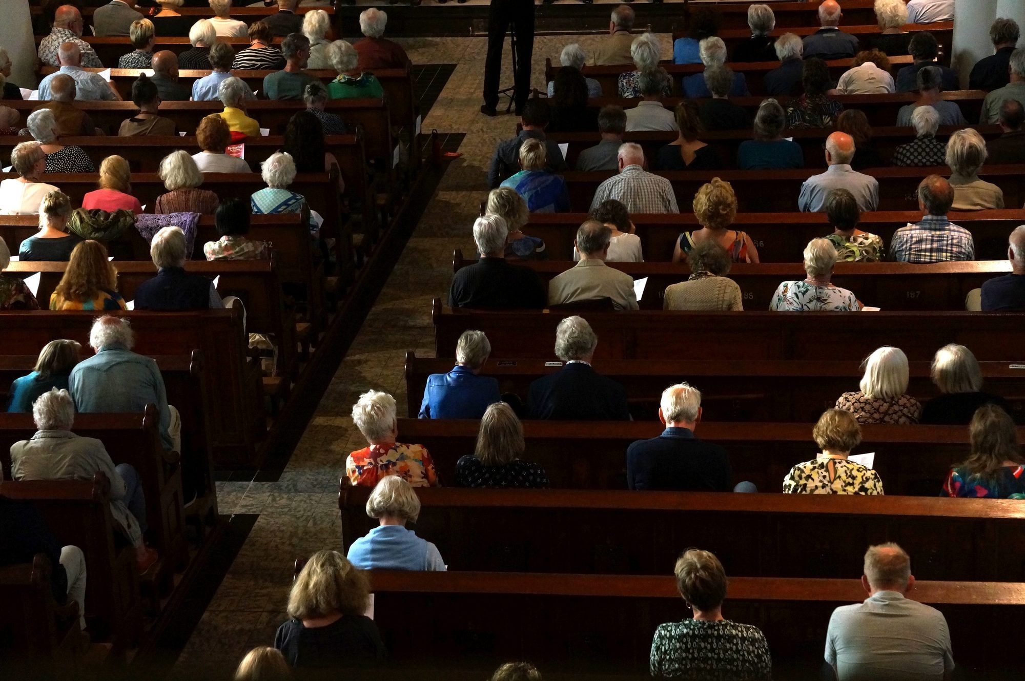 Rows of a congregation seated in wooden pews seen from above, service sheets in hand, a speaker at the front