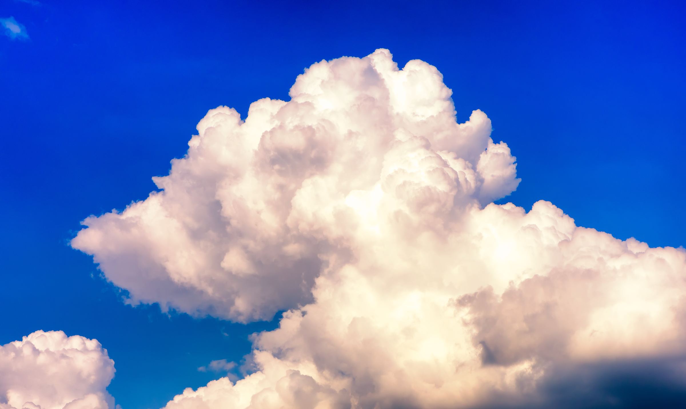 A towering white cumulus cloud, sunlit on its upper edges and shaded underneath, rising against a deep saturated blue sky