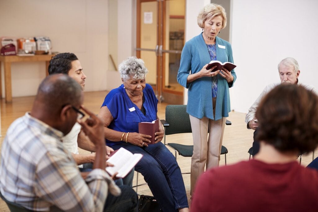 Six adults of different ages sit in a loose circle of chairs in a bright parish hall, several holding small red books, while a woman in a blue cardigan stands and reads aloud