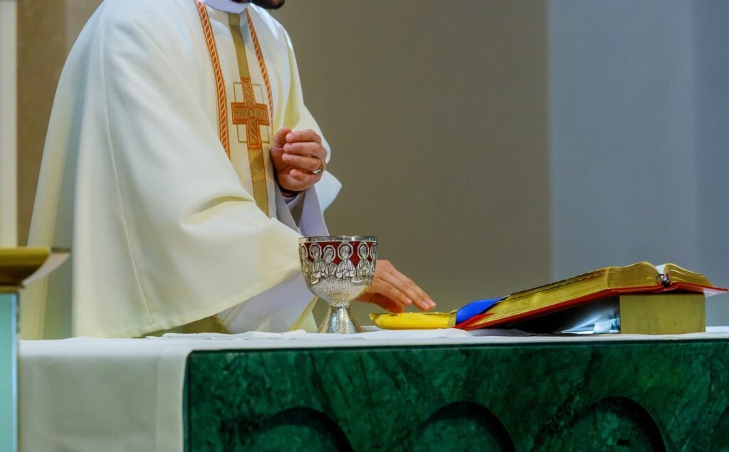 A priest in white vestments at the altar with a silver chalice and an open missal, a green altar frontal below