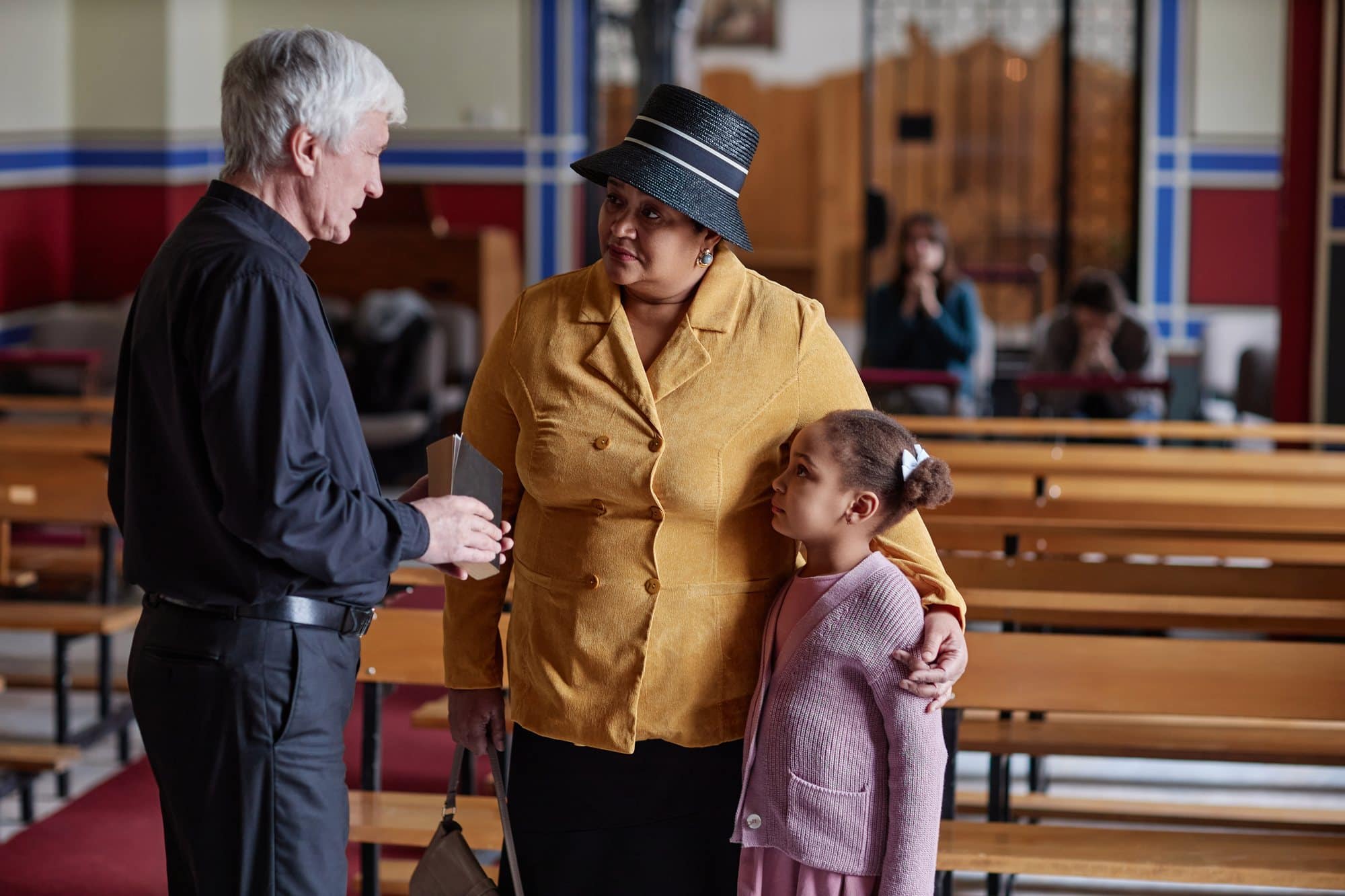 A grey-haired priest in black clerical dress talks with a woman in a mustard coat and black straw hat, her arm around a young girl in a pink cardigan, beside wooden pews in a church