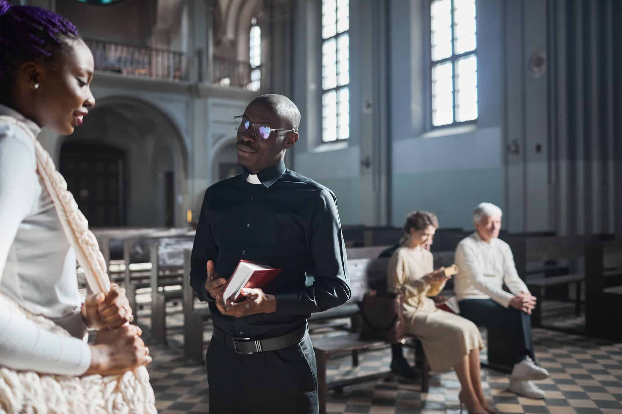 A priest in a black clerical shirt holds a red book and talks with a young woman in a white knitted top, standing in the aisle of a light-filled church while people sit in the pews behind