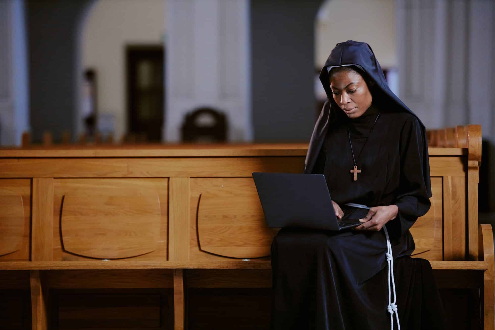 A religious sister in a black habit and veil with a wooden cross sits at the end of a wooden pew, typing on a laptop balanced on her knees, in a quiet church