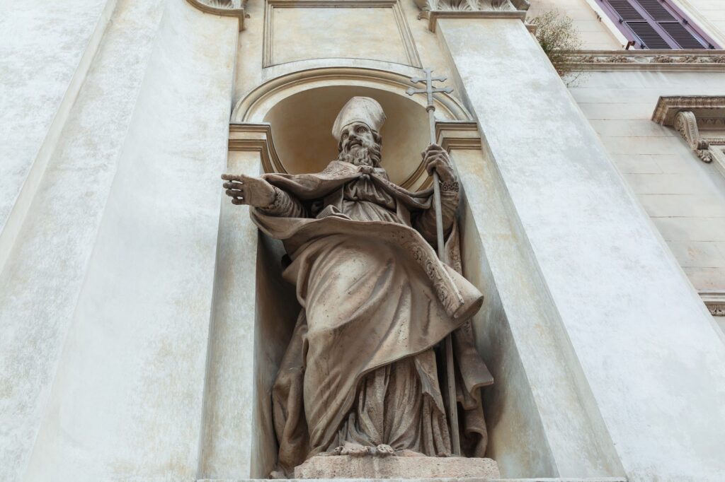 A stone statue of a bishop saint with a mitre and crozier in a niche on a church facade in Rome, one hand raised