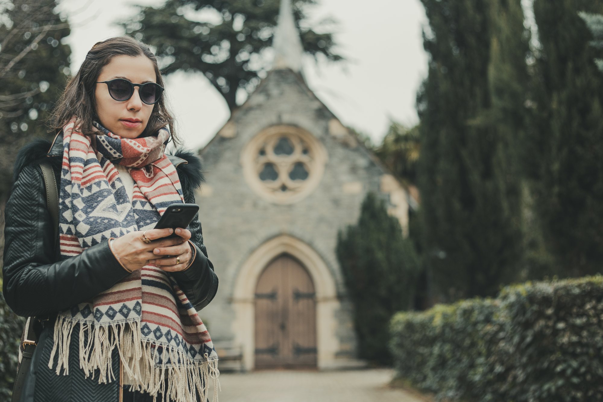 A woman in sunglasses, a black jacket and a patterned scarf looks down at her phone in front of a small stone chapel with an arched wooden door and a rose window