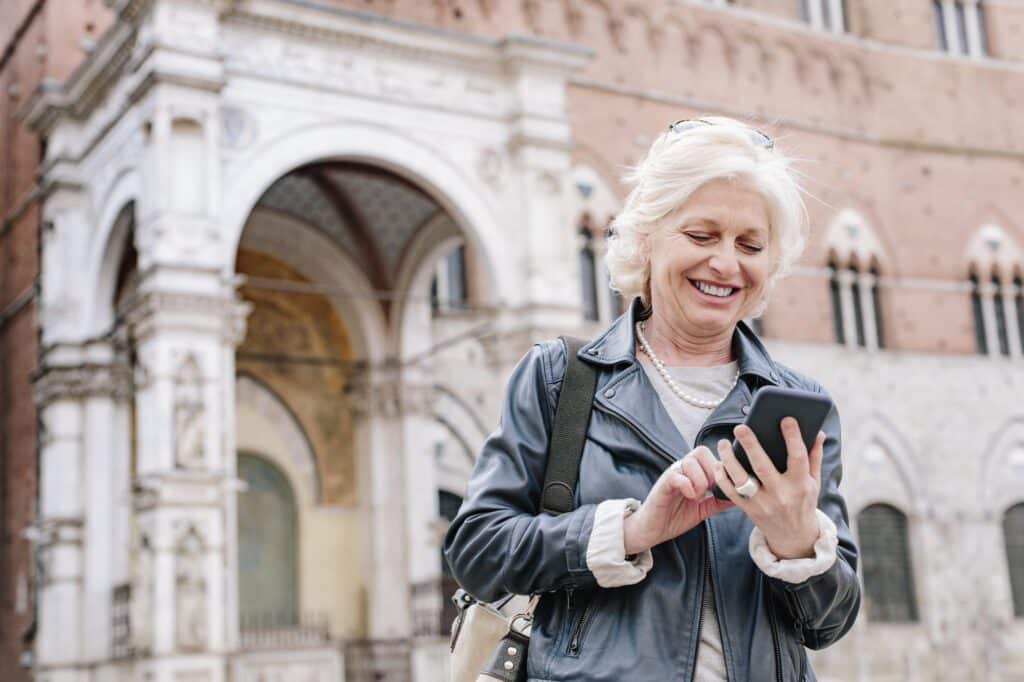 A woman with white hair in a leather jacket smiles down at her phone in a sunlit square, with the arches and rose-coloured stone of an old Italian church behind her